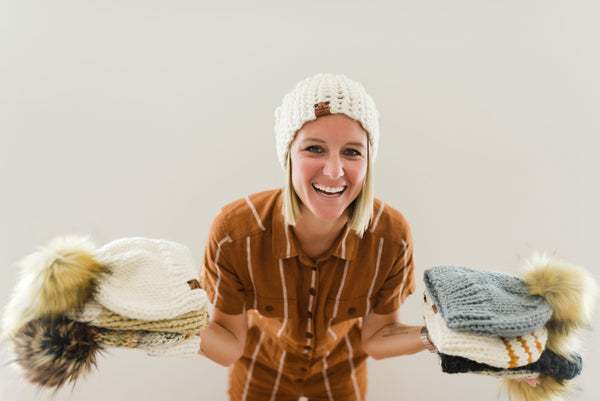 Brooke Happe holding stack of handmade knit winter pom pom hats