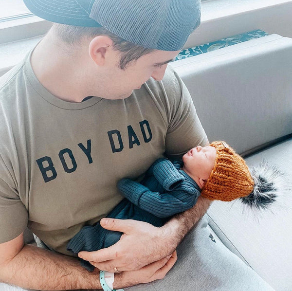 New father in a boy dad tee holding newborn son wearing a b.e.happe handmade knit pom pom hat