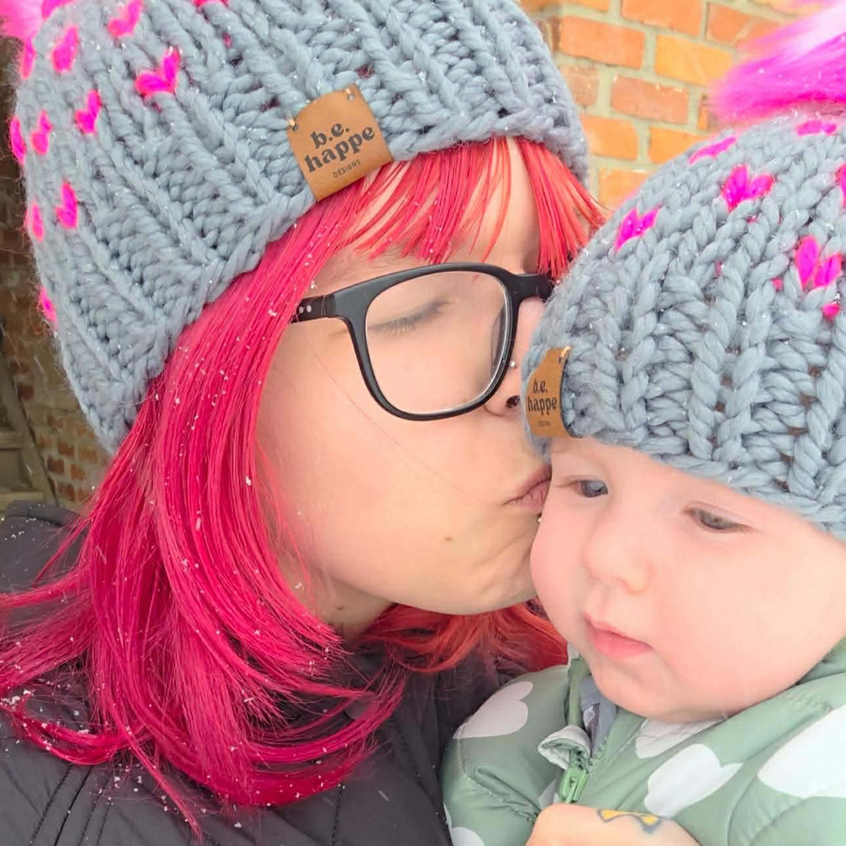 Person with pink hair and glasses kissing a child on the cheek, both wearing behappe mommy and me matching knitted hats.