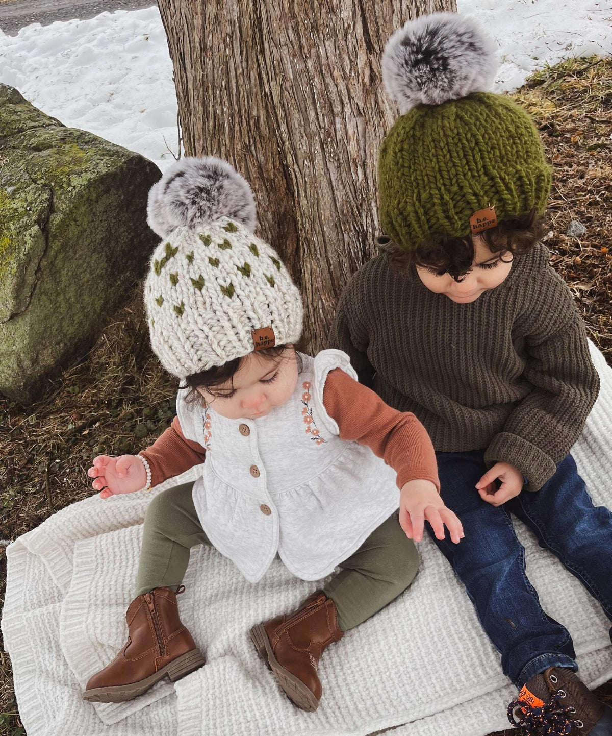 Two children wearing behappe matching knitted hats sitting on a blanket next to a tree.