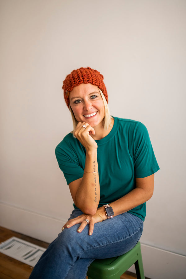 Brooke Happe wearing a green shirt and behappe orange knit slouch hat sitting on a green stool against a plain wall.