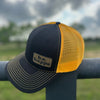 A black and yellow snapback trucker cap with a behappe brand patch on the bottom corner, placed on a metal railing with a cloudy sky in the background.
