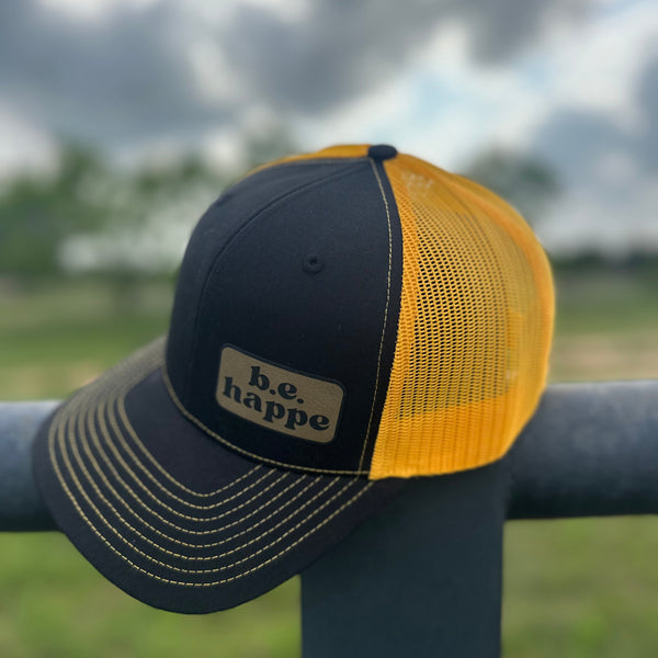 A black and yellow snapback trucker cap with a behappe brand patch on the bottom corner, placed on a metal railing with a cloudy sky in the background.