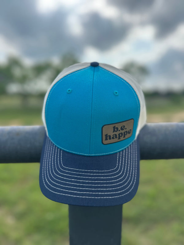 A blue trucker snapback cap with a white and black camo design visor, featuring a b.e.happe branded patch on the front.
