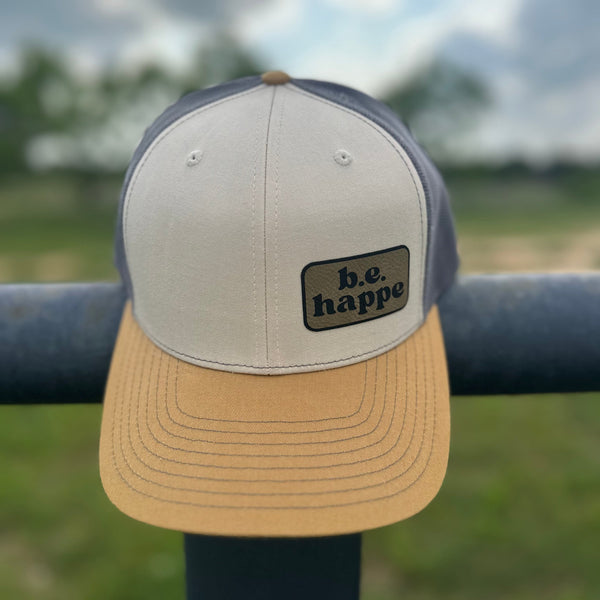 A snapback trucker cap in grey, cream, and muted mustard yellow colors, with a b.e.happe branded patch on the front.