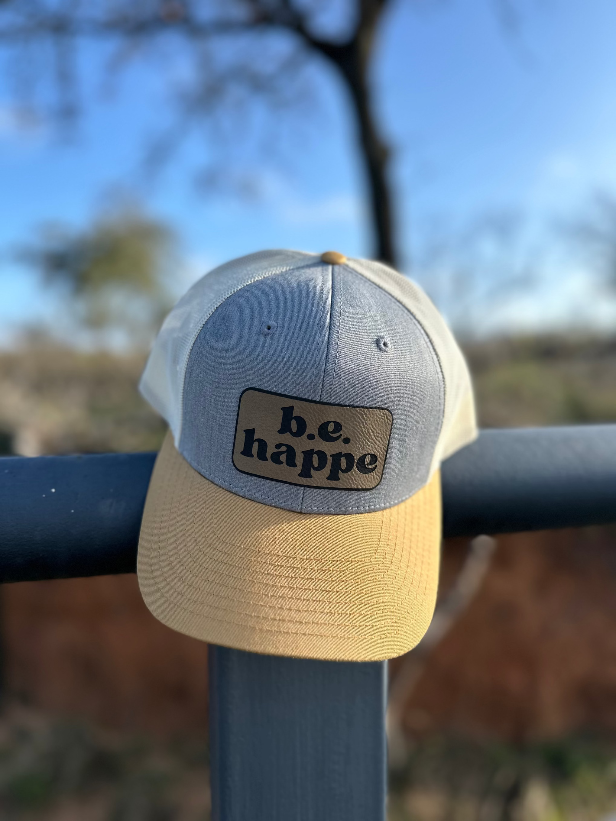 A trucker snapback cap with a camouflage design in gray and yellow, featuring a pre-curved visor with contrast stitching, and an adjustable plastic snapback strap. Tbehappe