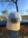 A trucker snapback cap with a camouflage design in gray and yellow, featuring a pre-curved visor with contrast stitching, and an adjustable plastic snapback strap. Tbehappe