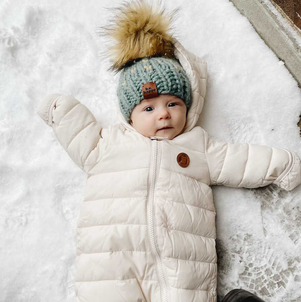 Baby in a white snowsuit and blue knit hat with pom-pom standing in the snow.