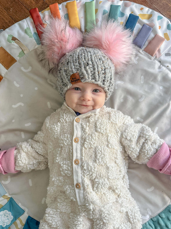 Baby wearing a behappe knitted hat with two pink pom-poms on a colorful play mat