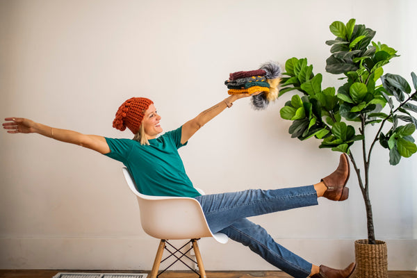 Brooke Happe sitting in a chair with arms outstretched, holding colorful handmade slouchy beanies and pom hats, next to a potted plant.