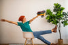 Brooke Happe sitting in a chair with arms outstretched, holding colorful handmade slouchy beanies and pom hats, next to a potted plant.