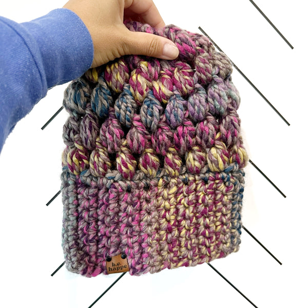 A multicolored crochet slouch hat being held by a hand, featuring a puff stitch design in purple and gray tones.