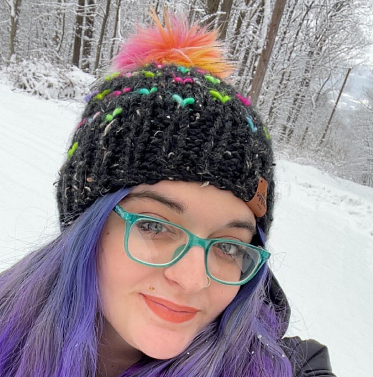 Person with colorful hair and a behappe rainbow pom beanie standing in a snowy landscape