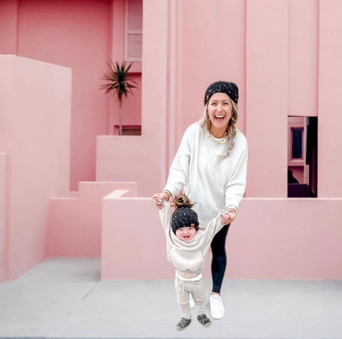 Woman and child in matching black beanies standing in front of a pink architectural background