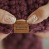 Close-up of a behappe purple knitted item with a beanie care  label held between fingers.