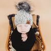 Newborn baby in a wooden basket wearing a behappe knitted hat with a pom-pom on a beige background