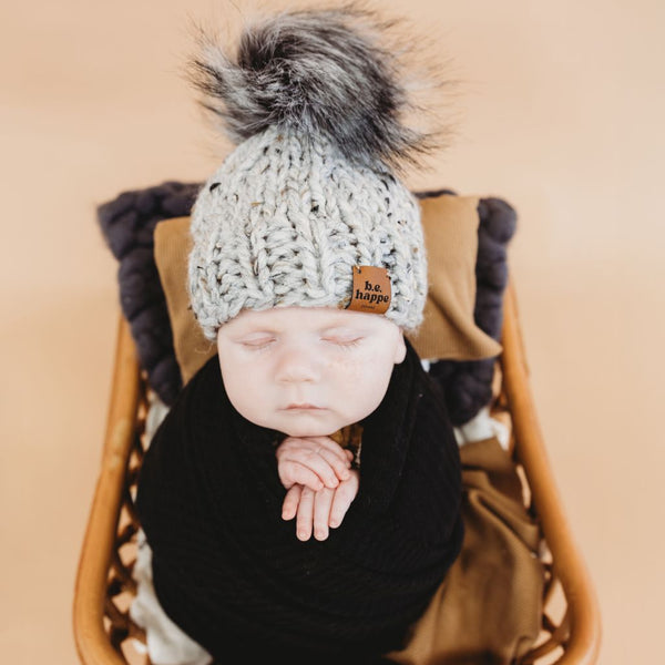 Newborn baby in a wooden basket wearing a behappe knitted hat with a pom-pom on a beige background