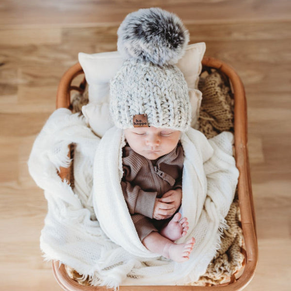 Newborn baby wrapped in a white blanket and wearing a behappe knitted hat with a pom-pom, lying in a wooden basket on a wooden floor.