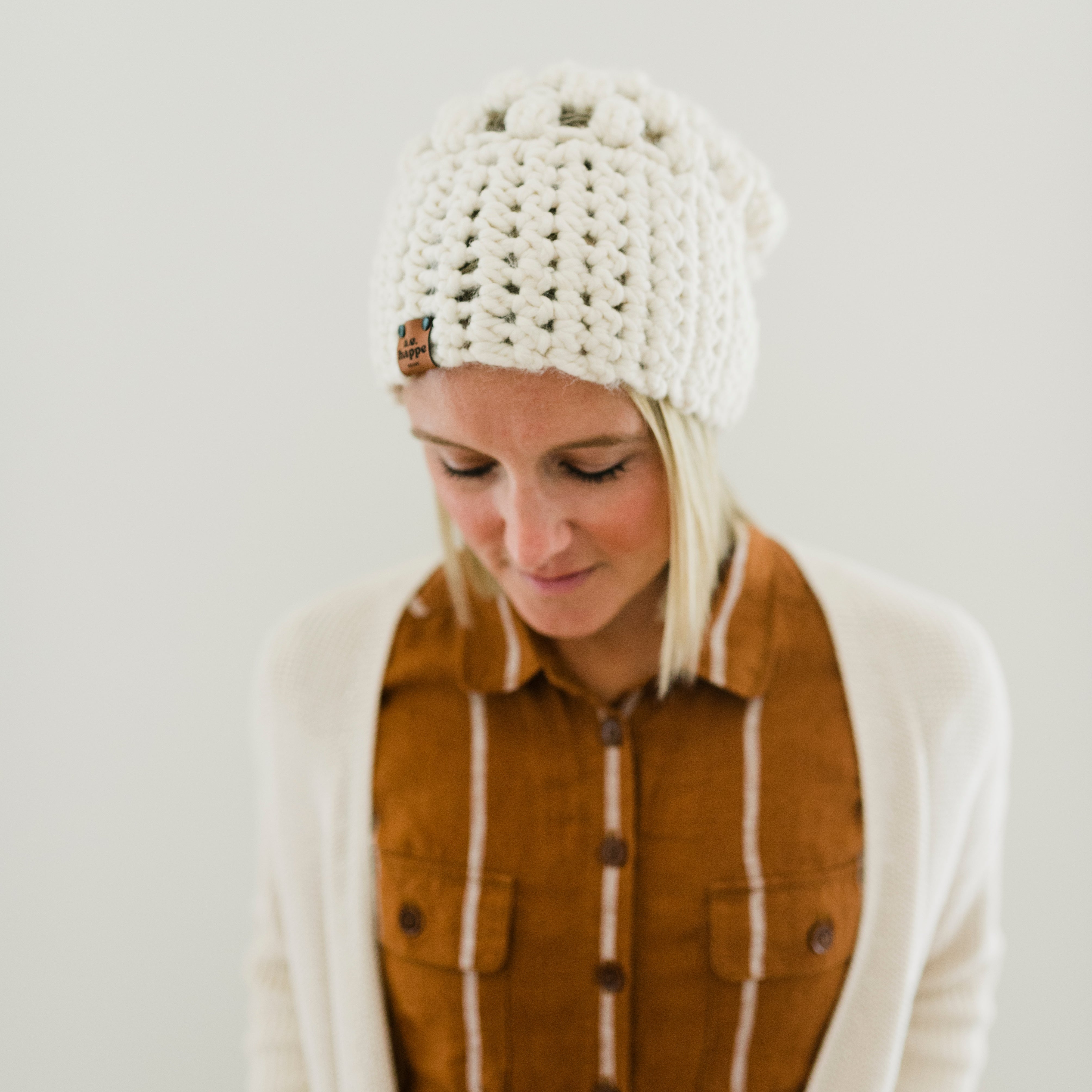 Woman wearing a white knitted beanie and brown jacket against a plain background