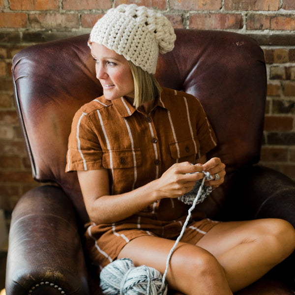 Woman sitting on a brown leather chair knitting with yarn and a wooden paddle, against a brick wall.
