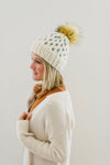 adult womens knit winter hat in off white with grey knit mini heart design and faux fur pom pom; handmade knit winter beanie with pom pom; faux fur winter pom pom hat; handmade knit winter stocking cap; womens stocking cap with pom pom; adult chunky winter beanie; handmade chunky knit heart hat; profile view of handmade knit winter hat with pom pom